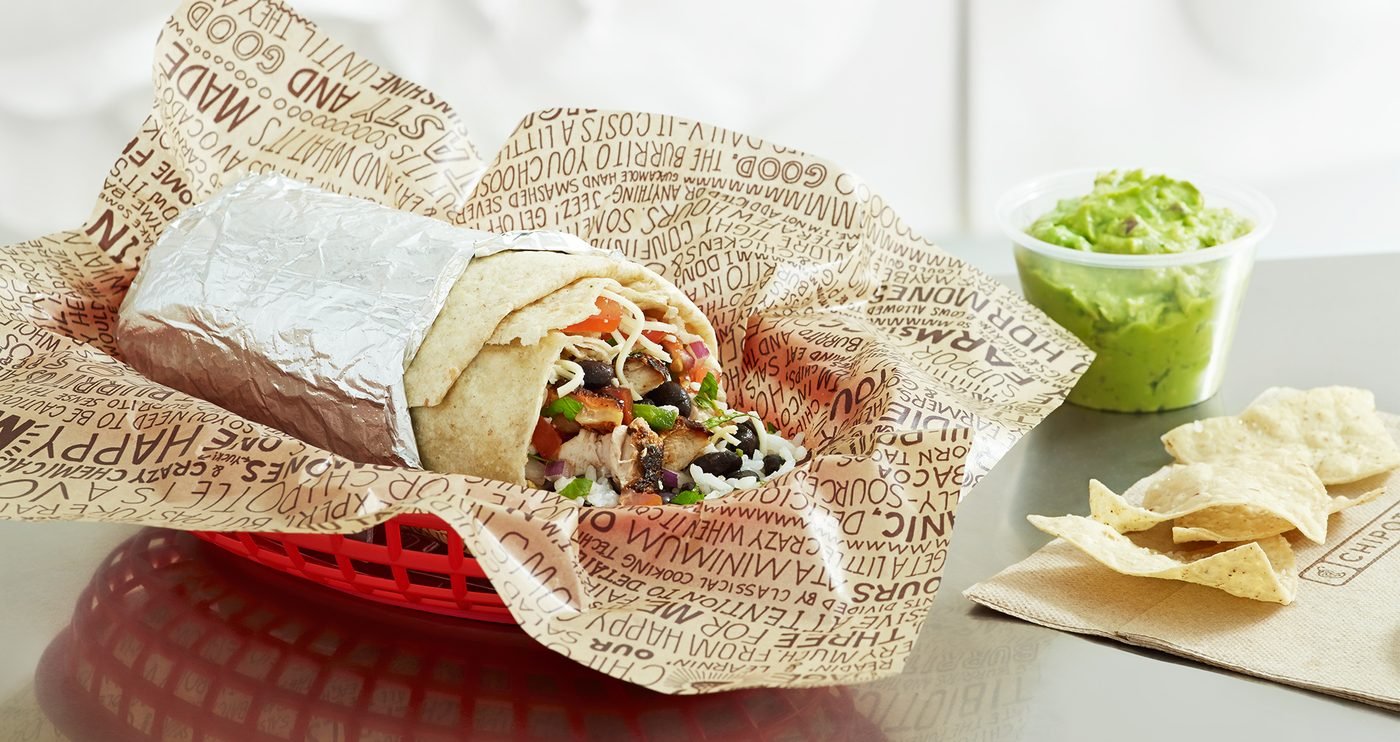 A foil-wrapped Chipotle burrito in a branded paper-lined basket with chips and guacamole