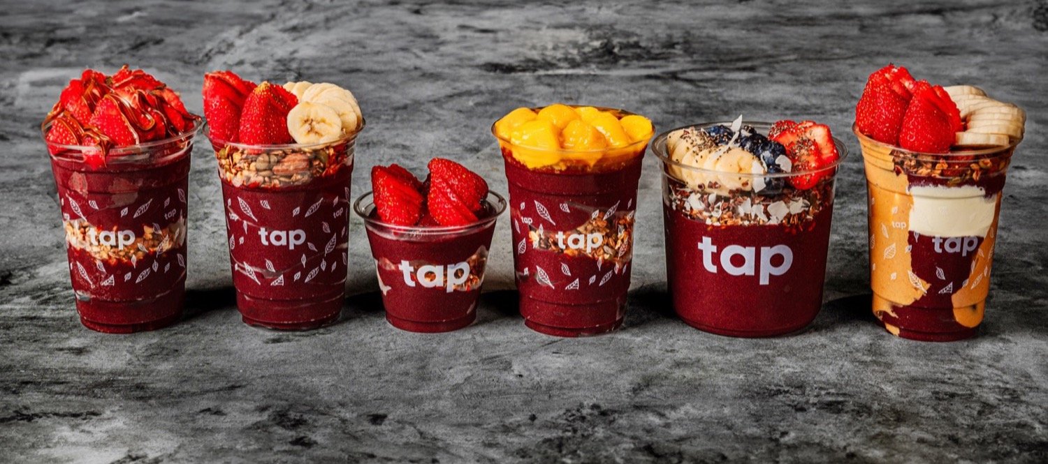 Six TAP açaí bowls in branded cups, topped with strawberries, bananas, mango, granola and Nutella